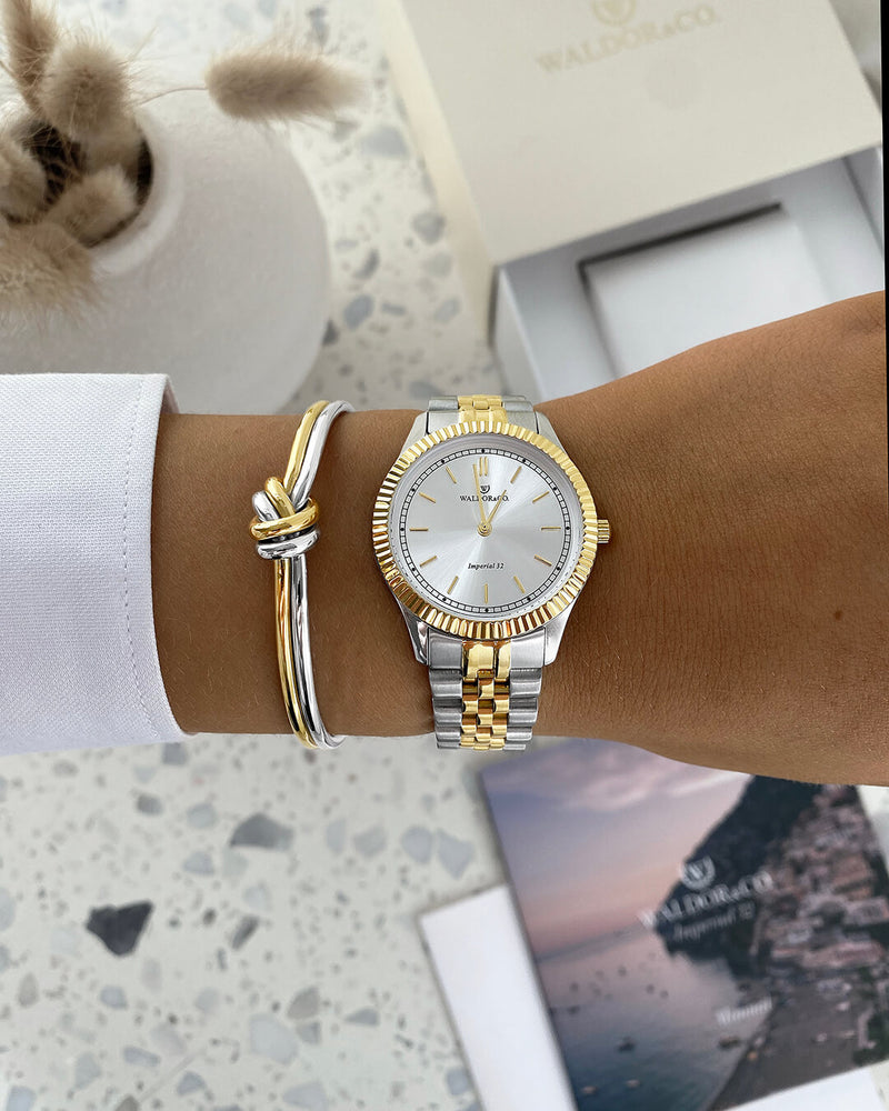 A round womens watch in silver and 22k gold from Waldor & Co. with silver sunray dial and a second hand. Seiko movement. The model is Imperial 32 Positano 32mm.
